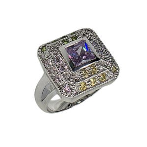 May include: A silver-toned ring with a square, multi-faceted, light purple center stone. The ring features a square setting with smaller, surrounding stones in shades of pink, green, and yellow. The ring's band is a smooth, polished silver.