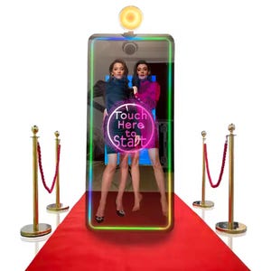 May include: A photo booth with a black frame and a colorful neon border. Two people are standing inside the booth, posing for a photo. The booth has a touch screen with the words "Touch Here to Start" displayed on it. The booth is set up on a red carpet with gold stanchions on either side.