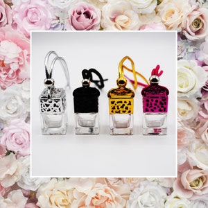 May include: Four glass car air fresheners with decorative tops in silver, black, gold, and magenta. Each has a small glass container and a hanging cord. The background features a floral arrangement of pink and white roses.