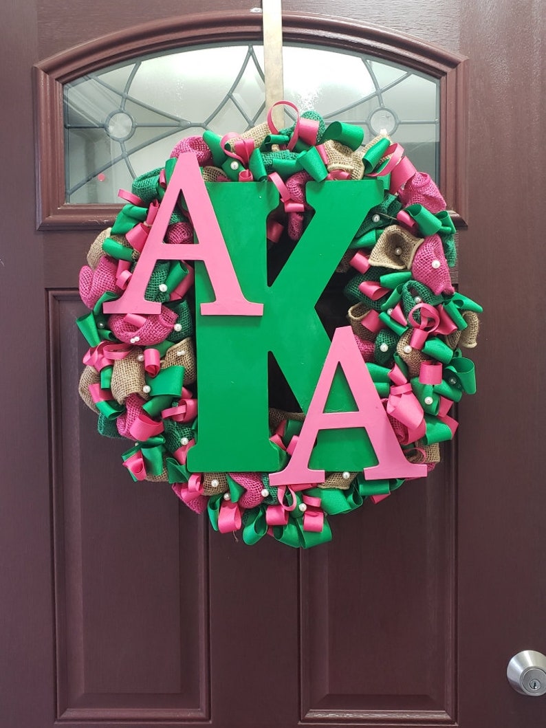 May include: A green and pink wreath with the letters "AKA" in pink on a green background. The wreath is made of burlap, ribbon, and other decorative elements.