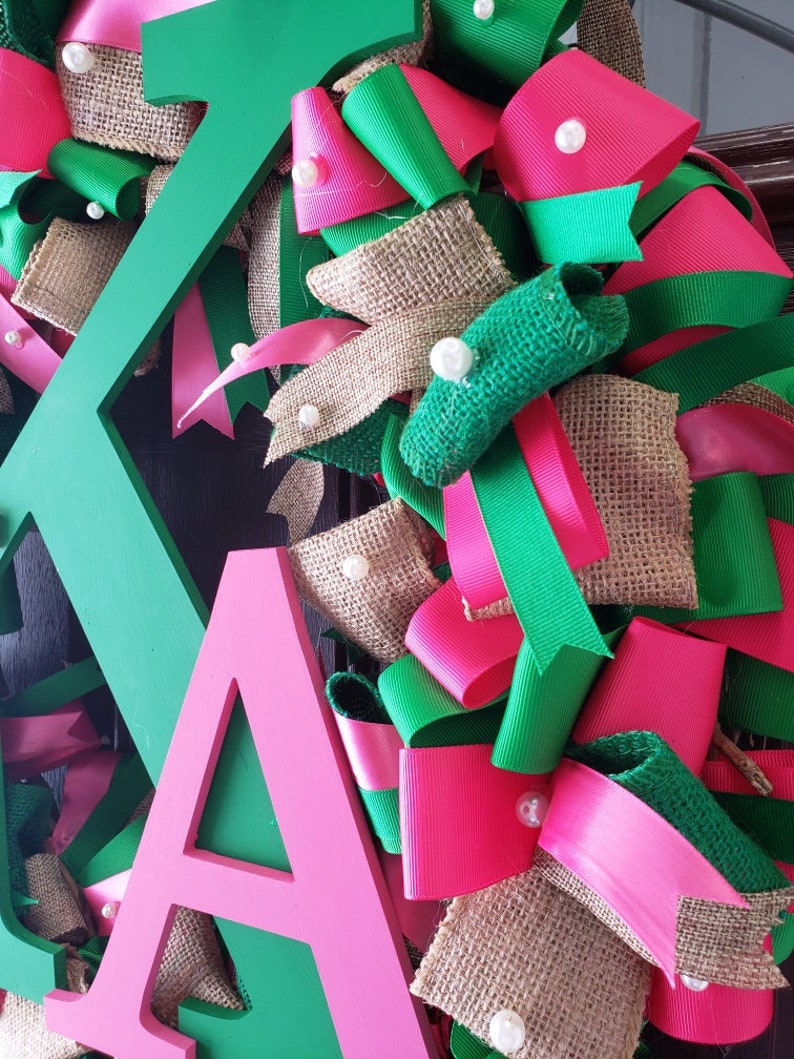 May include: A green and pink wreath with the letters "KA" in the center. The wreath is made of burlap, ribbon, and pearls.
