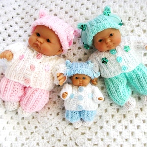 May include: Three knitted baby dolls wearing matching hats and outfits. The dolls are lying on a white crocheted blanket. The dolls are wearing pink, blue, and green outfits.