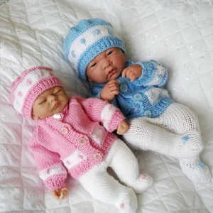 Könnte beinhalten: Zwei gestrickte Baby-Puppenoutfits, eines rosa und eines blau, mit passenden Mützen. Die Outfits haben weiße Akzente und Knopfverschlüsse. Die Puppen liegen auf einer weißen Decke.