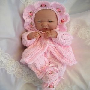 Può includere: Un completo per neonato in maglia rosa con un berretto abbinato. Il berretto ha un bordo a festoni ed è decorato con fiori rosa. Il completo ha una giacca stile cardigan e pantaloni abbinati.
