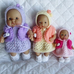 May include: Three knitted doll outfits in pink, yellow, and purple. Each outfit includes a dress, a cardigan, and a hat. The dolls are not included.