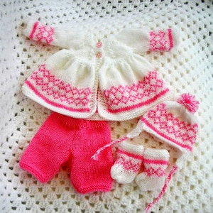Könnte beinhalten: Ein gestricktes Baby-Outfit in Rosa und Weiß. Das Set beinhaltet eine Strickjacke, eine Hose, eine Mütze und Booties. Die Strickjacke hat eine weiße Basis mit rosa Rautenmustern und einem rosa Saum. Die Hose ist einfarbig rosa. Die Mütze ist weiß mit rosa Rautenmustern und einem rosa Bommel. Die Booties sind weiß mit rosa Rautenmustern.