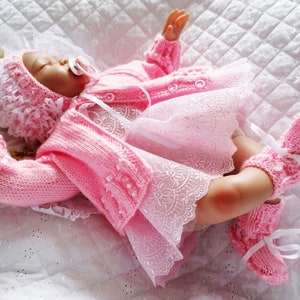 May include: A pink knitted baby outfit with a matching hat and booties. The outfit features a cardigan with white buttons and a lace trim skirt.
