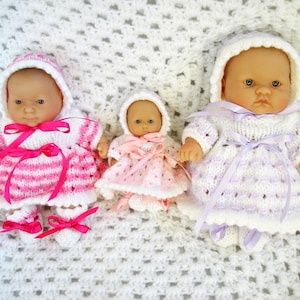 May include: Three knitted doll outfits in pink, white, and lavender. The dolls are wearing hats and have bows on their dresses.