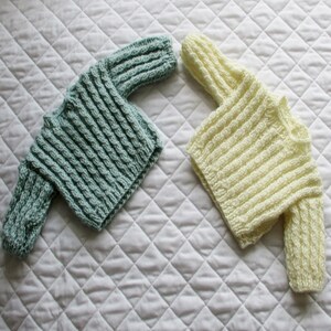 May include: Two knitted baby sweaters, one in a light blue color and the other in a cream color. Both sweaters have a cable knit pattern.