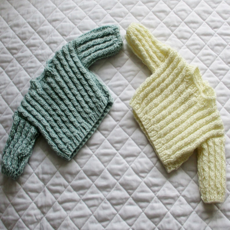 May include: Two knitted baby sweaters, one in a light blue color and the other in a cream color. Both sweaters have a cable knit pattern.