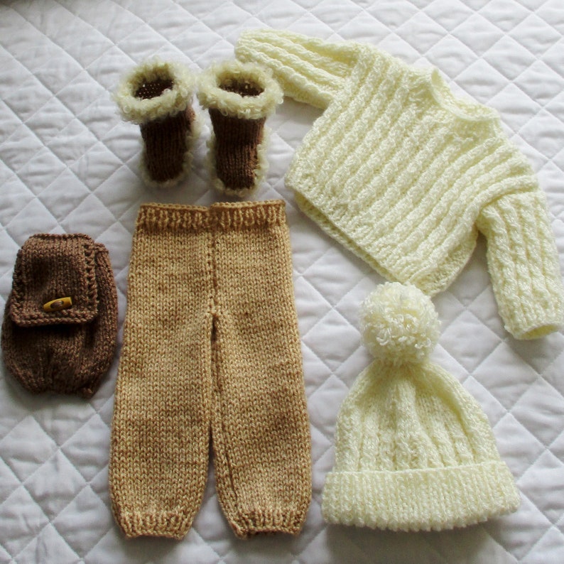 May include: A knitted baby outfit set in cream and brown. The set includes a sweater, pants, hat, and booties. The sweater has a ribbed texture and the pants have a smooth texture. The hat has a pom-pom on top. The booties have a faux fur trim.