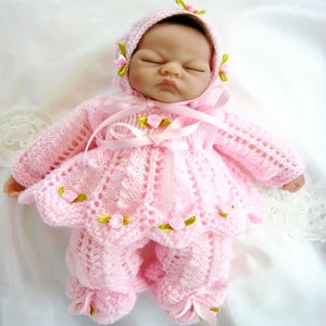 Puede incluir: Un conjunto de muñeca de bebé de punto rosa con un gorro a juego, con una cinta y adornos florales.