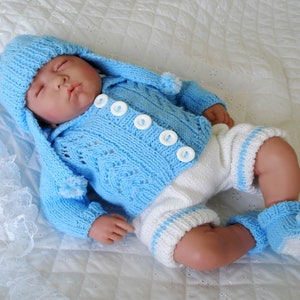 Op de afbeelding: Een blauwe gebreide baby-outfit met een bijpassende muts. De outfit bestaat uit een cardigan met witte knopen en een broek met witte strepen.