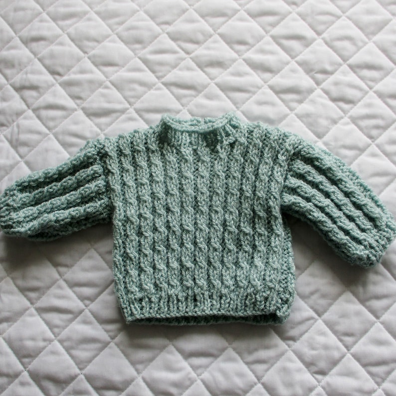 May include: A light green knitted baby sweater with a cable knit pattern.