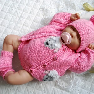 Könnte beinhalten: Ein rosa gestricktes Baby-Outfit mit passender Mütze und Stiefeln. Das Outfit hat ein kleines Bärenmotiv auf der Vorderseite. Das Baby trägt einen Schnuller.