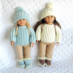 Op de afbeelding: Twee poppen die winterse gebreide outfits dragen. De pop links draagt een lichtblauwe trui, een beige broek en blauwe laarzen. De pop rechts draagt een crèmekleurige trui, een beige broek en bruine laarzen. Beide poppen dragen gebreide mutsen met pompoenen.