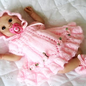 May include: A pink knitted baby doll dress with a ruffled neckline and matching booties. The dress has several small pink flowers sewn on it. The doll is wearing a pink dummy.