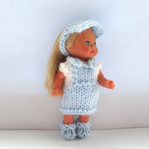 May include: A knitted doll outfit in light blue with a matching hat and shoes. The outfit features a sleeveless overall dress with a white shirt underneath.