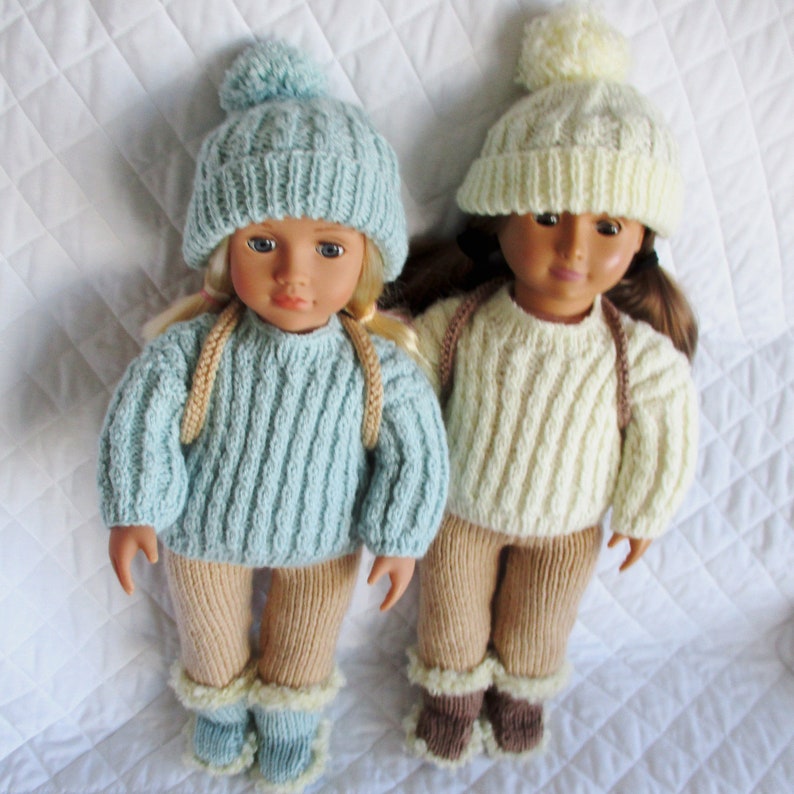 May include: Two dolls wearing knitted outfits. The doll on the left is wearing a light blue sweater, tan pants, and a light blue hat with a white pom-pom. The doll on the right is wearing a cream sweater, tan pants, and a cream hat with a white pom-pom.