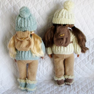 May include: Two knitted doll outfits with matching hats and backpacks. One outfit is light blue with a brown backpack, the other is white with a brown backpack.