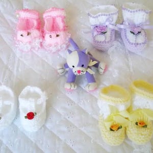 Pode incluir: Um conjunto de seis sapatinhos de bebé de crochê em rosa, branco e amarelo com detalhes florais. Os sapatinhos estão dispostos sobre uma superfície de crochê branca com um brinquedo de gato de crochê roxo.