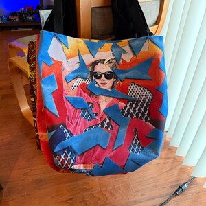 May include: A tote bag with a vibrant design featuring a woman wearing sunglasses, set against a backdrop of red, blue, and white geometric shapes. The bag has black straps and a colorful, eye-catching print.