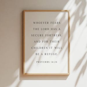 May include: A rectangular wall art print in a light wood frame featuring black serif text on a white background that reads: WHOEVER FEARS THE LORD HAS A SECURE FORTRESS, AND FOR THEIR CHILDREN IT WILL BE A REFUGE. PROVERBS 14:26.