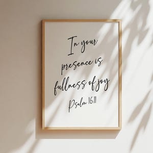 May include: A rectangular wall art print in a light wood frame featuring the black text In your presence is fullness of joy Psalm 16:11 written in a cursive script font on a white background, displayed against a plain neutral wall.