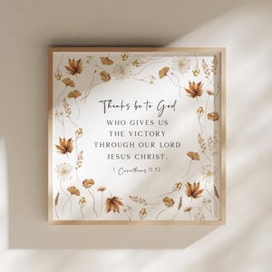 May include: A square framed wall art piece featuring a white background with a border of brown and tan autumn leaves and dried flowers. The center text reads: Thanks be to God WHO GIVES US THE VICTORY THROUGH OUR LORD JESUS CHRIST. 1 Corinthians 15:57.
