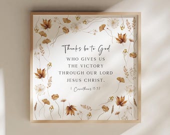 May include: A square framed wall art piece featuring a white background with a border of brown and tan autumn leaves and dried flowers. The center text reads: Thanks be to God WHO GIVES US THE VICTORY THROUGH OUR LORD JESUS CHRIST. 1 Corinthians 15:57.