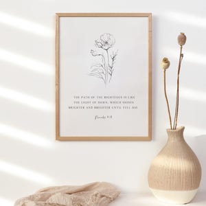 May include: A framed wall art print featuring a black line drawing of a flower above the text: THE PATH OF THE RIGHTEOUS IS LIKE THE LIGHT OF DAWN, WHICH SHINES BRIGHTER AND BRIGHTER UNTIL FULL DAY. Proverbs 4:18. The frame is light-colored wood.
