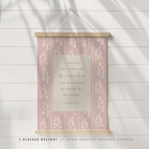 May include: A hanging canvas wall art piece featuring a dusty pink background with a white vertical leaf pattern. A central beige rectangle displays the text: I SOUGHT THE LORD, AND He answered me AND DELIVERED ME FROM ALL MY FEARS. PSALM 34:4.