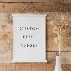 May include: A white rectangular wall hanging with a wooden frame at the top and bottom, featuring the black text CUSTOM BIBLE VERSES printed in the center. The piece is suspended by a thin string and displays a clean, minimalist aesthetic.