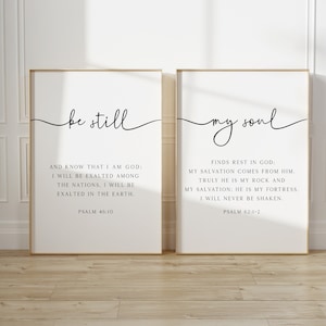 May include: Two vertical rectangular wall art prints in thin gold frames. The left print features the black text "be still" in script and "AND KNOW THAT I AM GOD: I WILL BE EXALTED AMONG THE NATIONS. I WILL BE EXALTED IN THE EARTH. PSALM 46:10". The right print features "my soul" in script and "FINDS REST IN GOD: MY SALVATION COMES FROM HIM. TRULY HE IS MY ROCK AND MY SALVATION: HE IS MY FORTRESS. I WILL NEVER BE SHAKEN. PSALM 62:1-2".