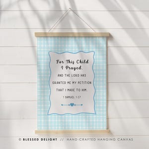May include: A hanging canvas wall art piece featuring a light blue and white gingham pattern background. The center displays a white scalloped-edge rectangle with the black text: For This Child I Prayed, AND THE LORD HAS GRANTED ME MY PETITION THAT I MADE TO HIM. 1 SAMUEL 1:27, with a small blue heart icon below.