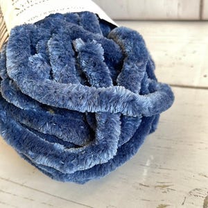 May include: A close-up of a ball of blue yarn. The yarn is thick and fluffy, with a soft, velvety texture. The yarn is a deep shade of blue, with lighter highlights. The yarn is made of polyester.