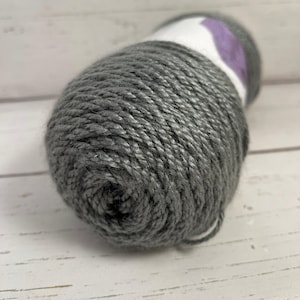 May include: A skein of gray yarn with a white label and a purple accent. The yarn is tightly wound, showing the texture of the fibers. Ideal for knitting or crocheting projects.