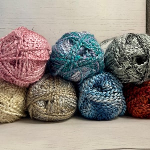 May include: Assortment of yarn balls in various colors, including pink, teal, silver, blue, and orange. The yarn is a sparkly, metallic type.
