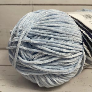 May include: A ball of soft, light blue yarn. The yarn has a velvety texture and is tightly wound. The label is partially visible, with a black and white striped pattern. Ideal for knitting or crocheting projects.