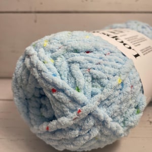 May include: A close-up of a ball of light blue yarn with colorful flecks. The yarn is thick and textured, with a label visible on the side. The yarn is for knitting and crocheting projects.