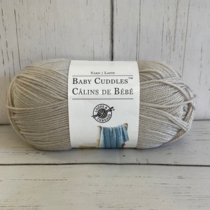 May include: A skein of Loops & Threads Baby Cuddle yarn in a light beige color. The label reads "Yarn | Laine Baby Cuddle Câlins de Bébé".