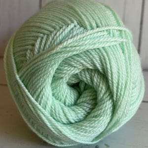 May include: A tightly wound ball of light green yarn. The yarn is composed of numerous strands, creating a textured surface. The color is a soft, pastel green, suitable for various knitting or crochet projects. The yarn appears soft and pliable.