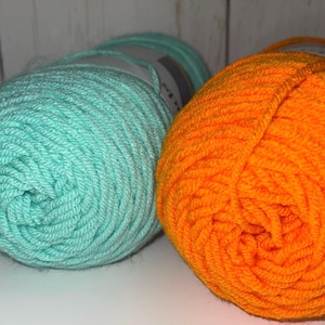 May include: Close-up of two yarn skeins. One is a vibrant turquoise, and the other is a bright orange. The yarn is tightly wound, showing the texture of the fibers. Ideal for knitting or crocheting projects.