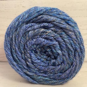 May include: A tightly wound ball of blue yarn with varying shades of light and dark blue, and hints of gray. The yarn appears thick and textured, suitable for knitting or crocheting projects. The yarn is on a white wooden surface.