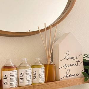 Puede incluir: Tres botellas de vidrio de mezclas de aceites esenciales para difusores con la marca "ISHA" y el texto "Natural Purity" en la etiqueta. Las botellas están dispuestas en un estante de madera con un letrero blanco que dice "home sweet home".