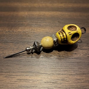 Könnte beinhalten: Dekorative Nadel in Schädelform mit gelbem Schädel, hellbrauner Perle und silberfarbenen Metallakzenten. Die Nadel hat eine lange, spitze Nadel und eine kleine Schlaufe am Ende des Schädels.