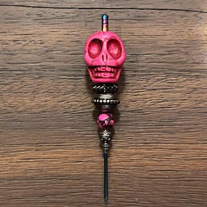 Könnte beinhalten: Ein dekoratives Objekt in Form eines rosa Schädels mit einer Metallspitze. Der Schädel hat dunkelrote Akzente und ist mit verschiedenen Metallperlen und Akzenten verziert. Die Spitze ist dunkelblau.