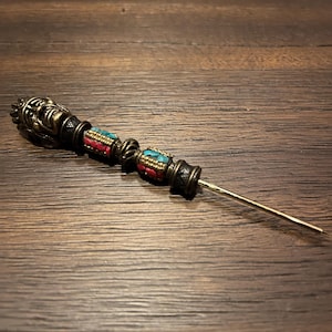Könnte beinhalten: Eine antike Nadel mit einem dekorativen Kopf mit einem Gesichtsdesign. Die Nadel hat einen messingfarbenen Schaft und ist mit roten und türkisfarbenen Perlen verziert. Die Nadel befindet sich vor einem dunklen Holzmaserungshintergrund.