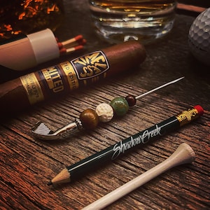 Könnte beinhalten: Ein Stillleben mit einer Zigarre, einem Golfball und einem Bleistift mit der Aufschrift "Shadow Creek". Ein Zigarrenpicker mit dekorativen Perlen und einem Golfschläger-förmigen Ende ist ebenfalls vorhanden. Die Gegenstände liegen auf einer Holzoberfläche.