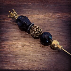 Könnte beinhalten: Ein dekorativer Flaschenverschluss mit schwarzem Totenkopf, goldener Krone, gold-schwarzen Perlen und einem goldenen Stiel. Der Verschluss ist für eine Weinflasche konzipiert. Der Hintergrund ist eine dunkle Holzoberfläche.
